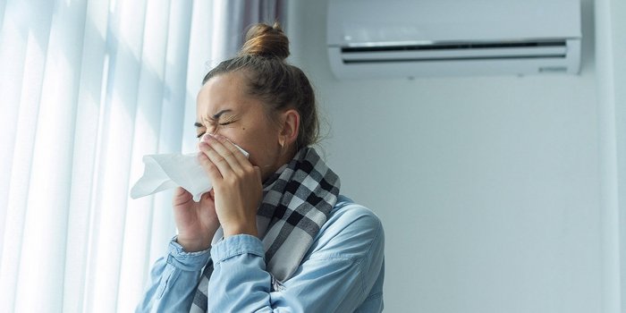 Im Vordergrung ist eine Frau in einem Raum, die Ihre Nase mit einem Taschentuch putzt. Im Hintergrund sieht man eine Klimaanlage an der Wand.